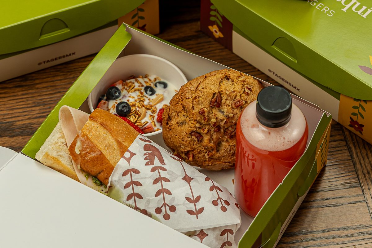 Box Lunch de Tagers abierto: baguette de carnes frías al pesto, galleta, jugo oasis y fruta con yogur; atrás, más cajas cerradas