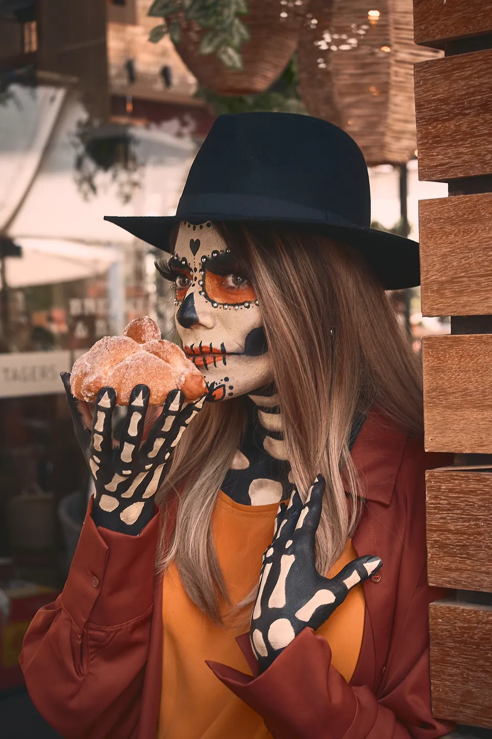 Una catrina de sombrero negro y guantes de esqueleto muerde un pan de muerto en la terraza de Tagers