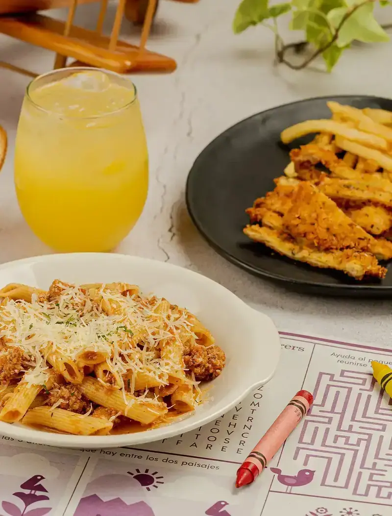Pasta a la boloñesa con queso, milanesa con papas y una naranjada sobre el mantel de juegos de Tagers, con crayolas