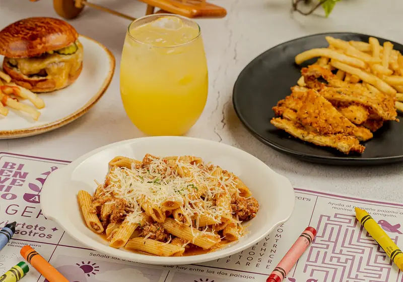 Mini smash burger con papas, pasta a la boloñesa, milanesa con papas y una naranjada sobre el mantel de juegos de Tagers, con crayolas