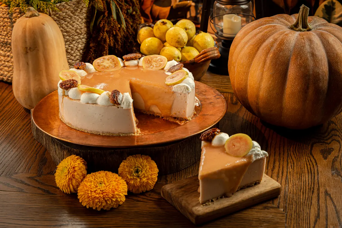 Cheesecake de guayaba familiar ya partido y una rebanada servida en su tabla, entre calabazas, guayabas y flores de cempasúchil