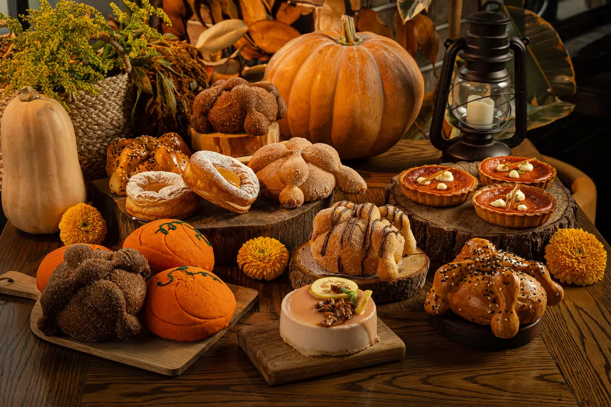 Mesa de roble con pan de muerto, conchas de calabaza, pays y cheesecake de temporada entre calabazas, flores de cempasúchil y un quinqué