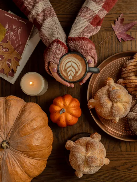 Manos con suéter sosteniendo un latte junto a pan de muerto, calabazas y una vela