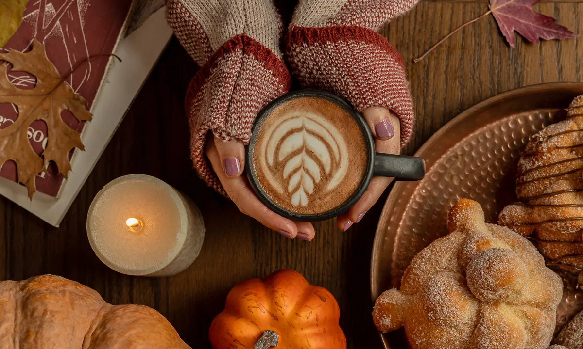 Manos con suéter sosteniendo un latte junto a pan de muerto, calabazas y una vela