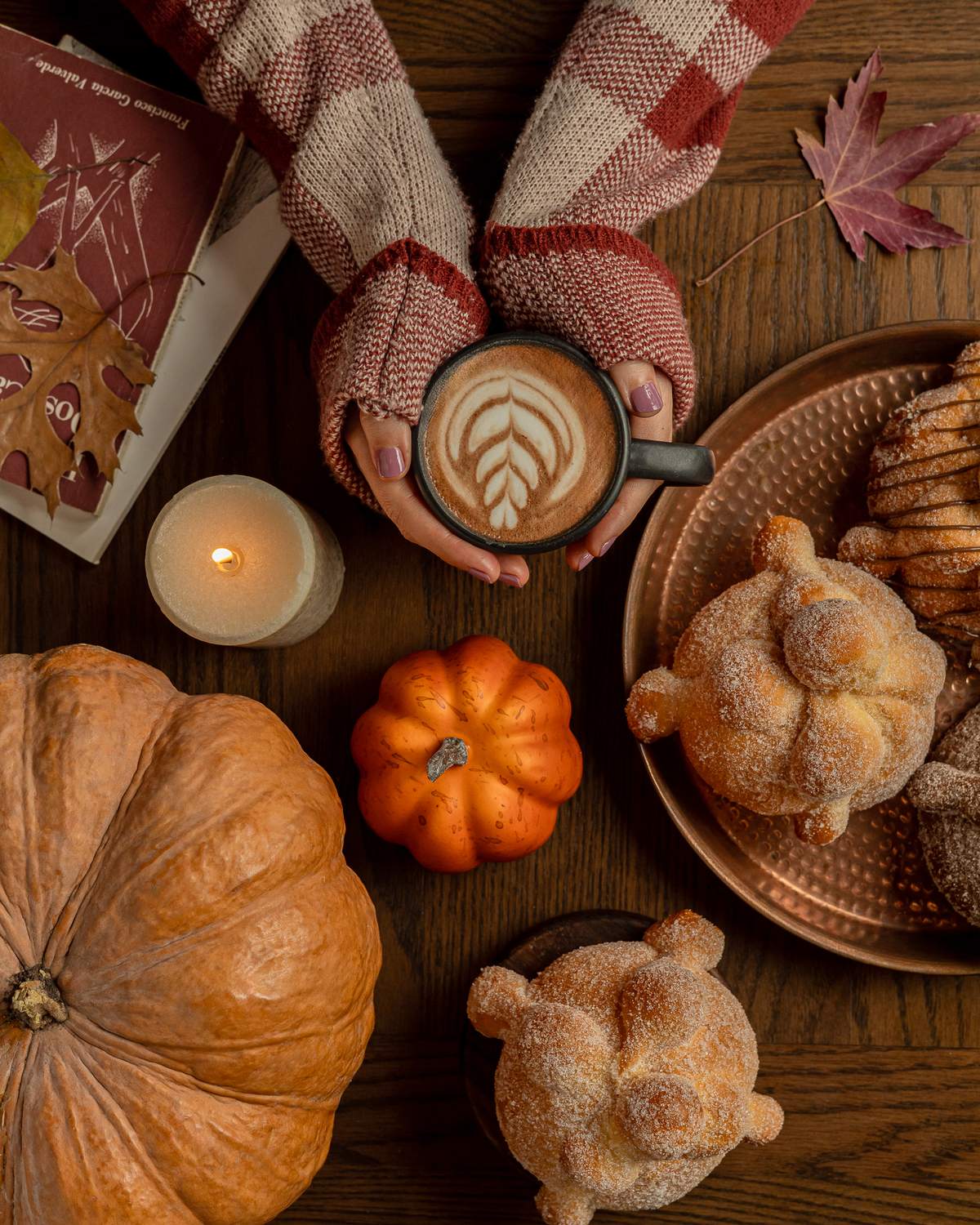 Manos con suéter sosteniendo un latte junto a pan de muerto, calabazas y una vela
