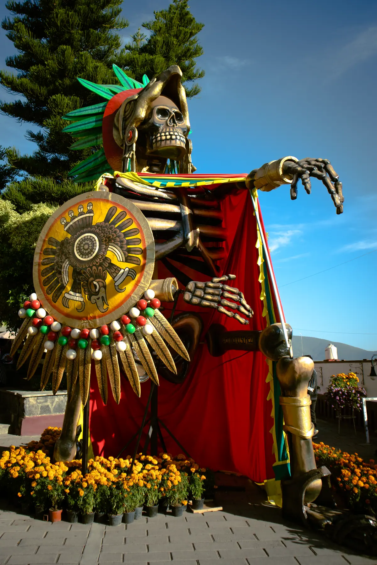 Catrina monumental de Cuauhtémoc con penacho, escudo y capa roja entre árboles, en el Valle de Catrinas de Atlixco en 2024