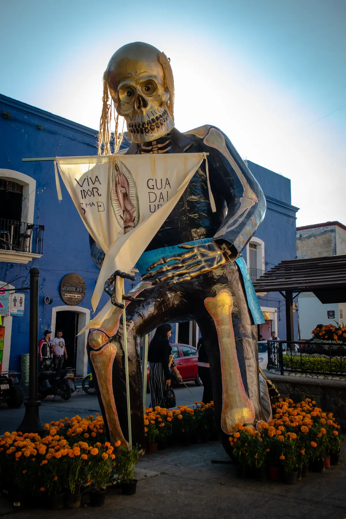 Catrina monumental de Miguel Hidalgo con un estandarte, frente a una casa azul del centro de Atlixco, en 2024