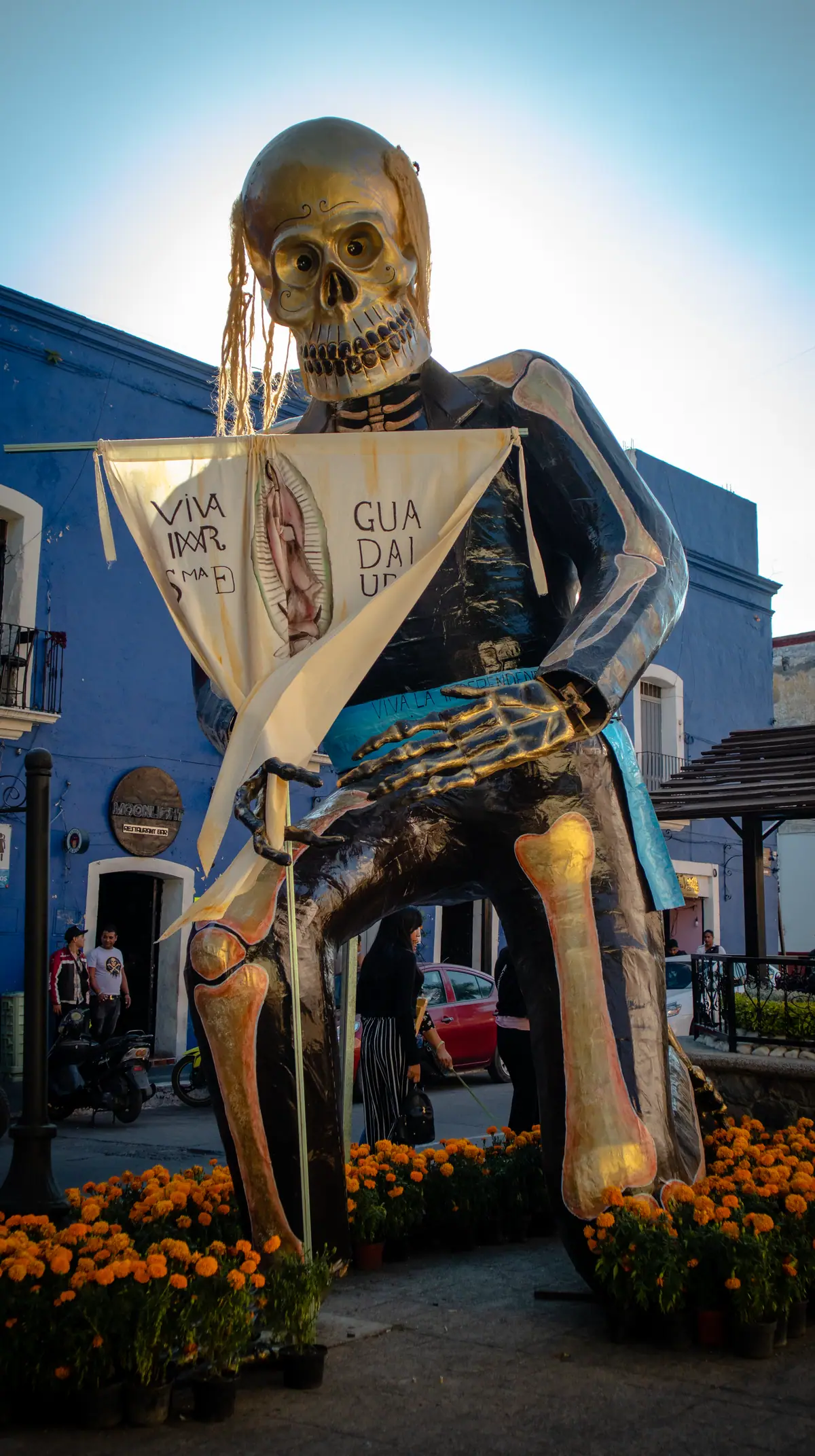 La catrina monumental de Miguel Hidalgo en una calle de Atlixco en 2024: de pie sobre el cempasúchil, con el estandarte de la Virgen de Guadalupe; abajo, entre sus piernas, pasa una mujer