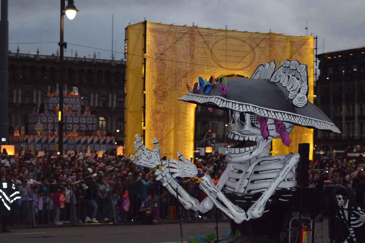 Esqueleto gigante con sombrero de catrina con plumas frente a un portal amarillo iluminado, con público detrás de las vallas, en el Gran Desfile de Día de Muertos de la Ciudad de México