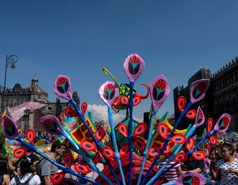 Un alebrije monumental con plumas en forma de flores entre el público en el Zócalo, con el Palacio Nacional al fondo