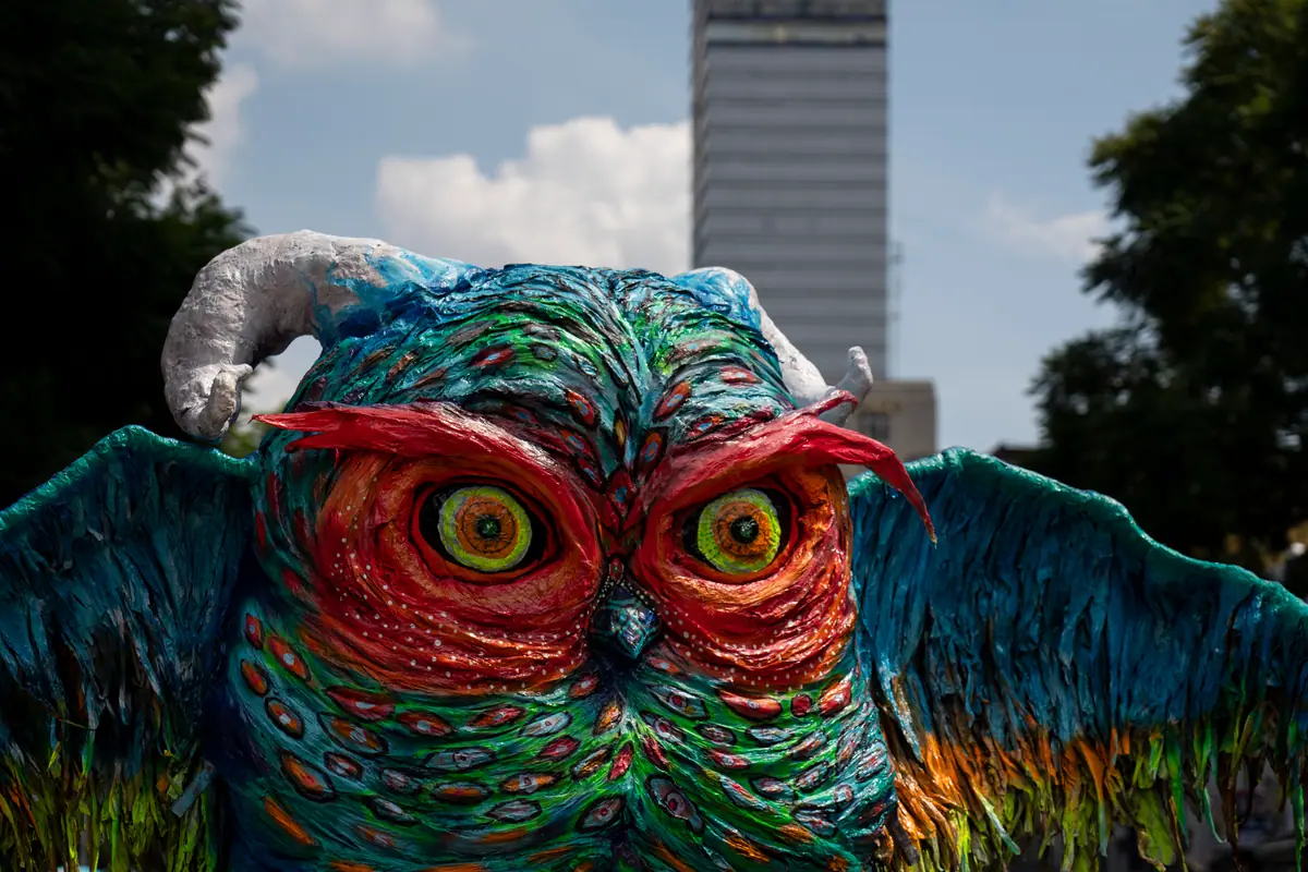 Alebrije monumental en forma de búho de colores, con una torre al fondo, en el desfile de alebrijes