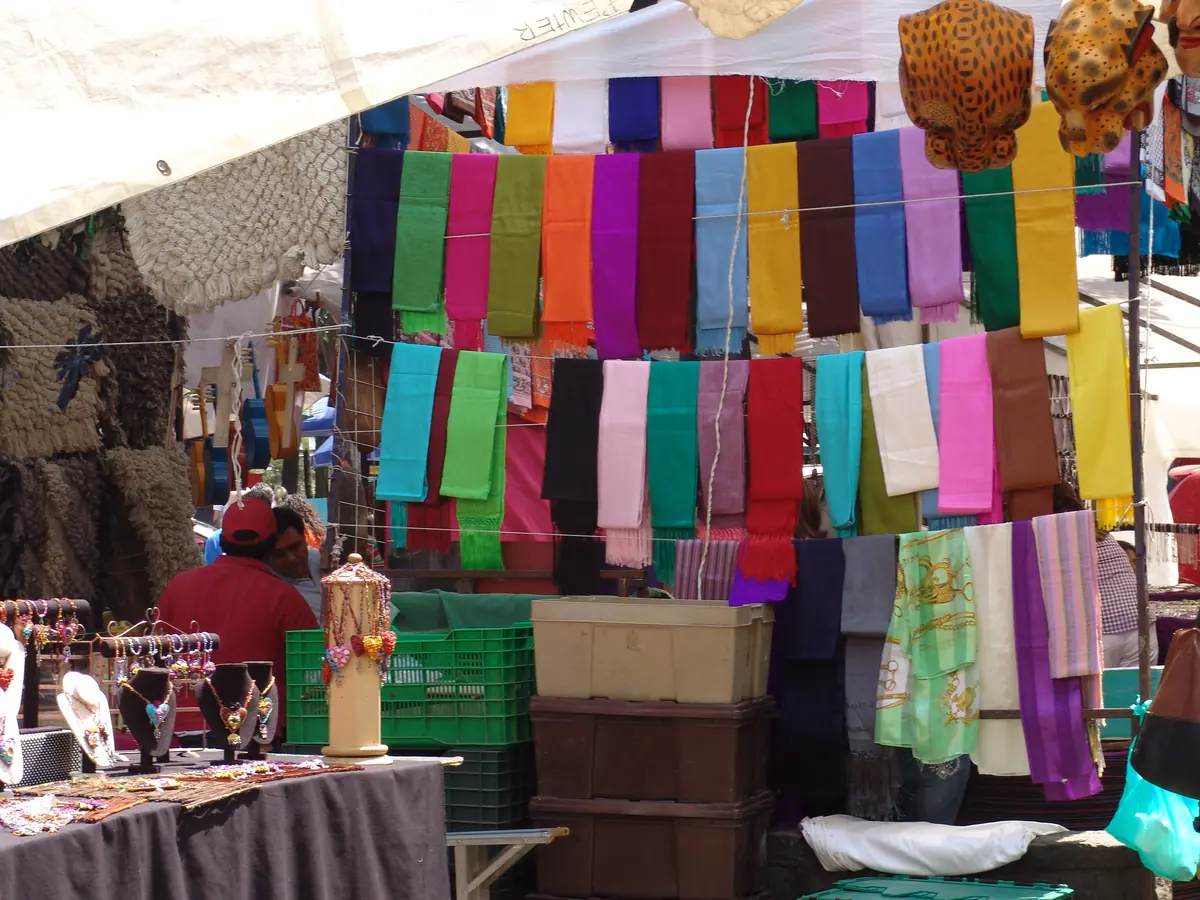 Un puesto bajo carpa en San Ángel con rebozos y chales de colores colgados en fila y bisutería sobre la mesa