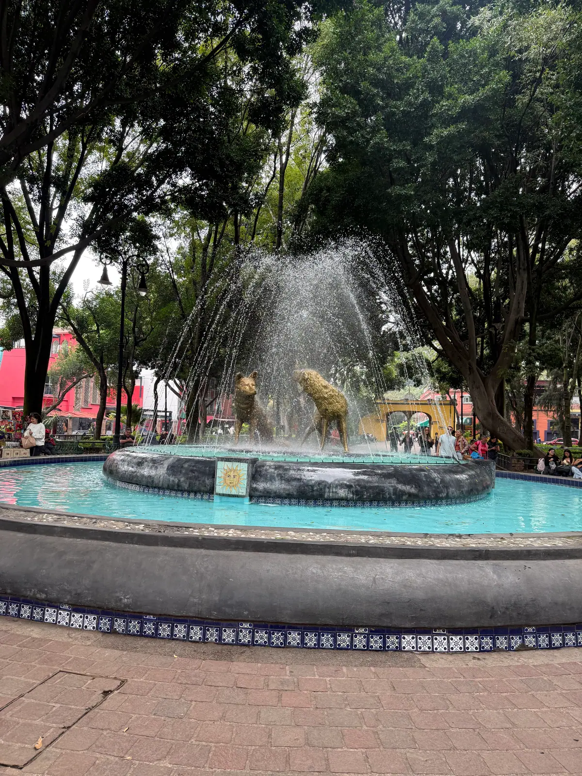 La Fuente de los Coyotes en el Jardín Centenario de Coyoacán: dos esculturas de coyotes entre chorros de agua y, al fondo, los arcos amarillos
