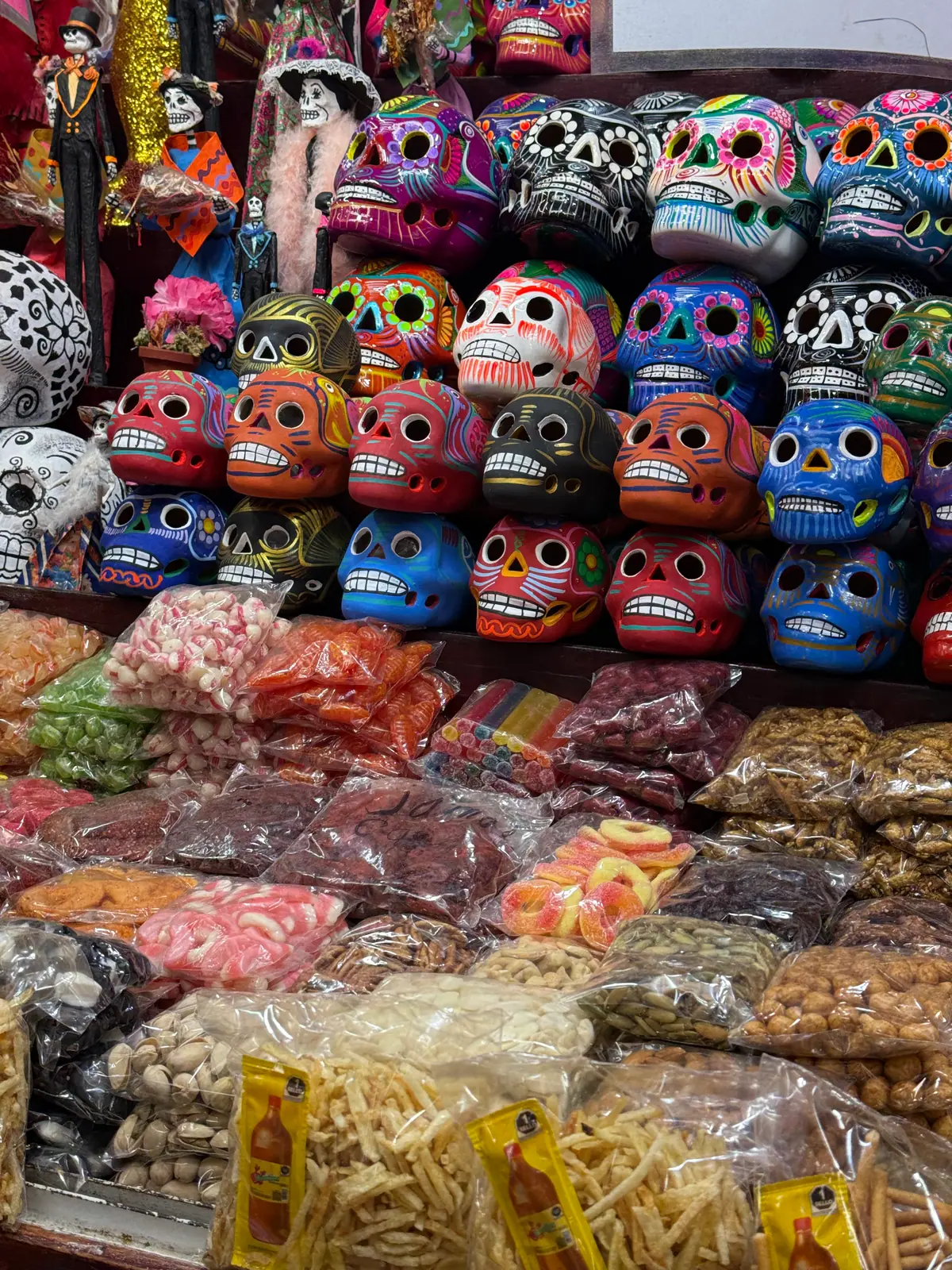 Calaveras de cerámica pintadas de azul, rojo, negro y morado apiladas sobre bolsas de dulces, gomitas, cacahuates y frituras