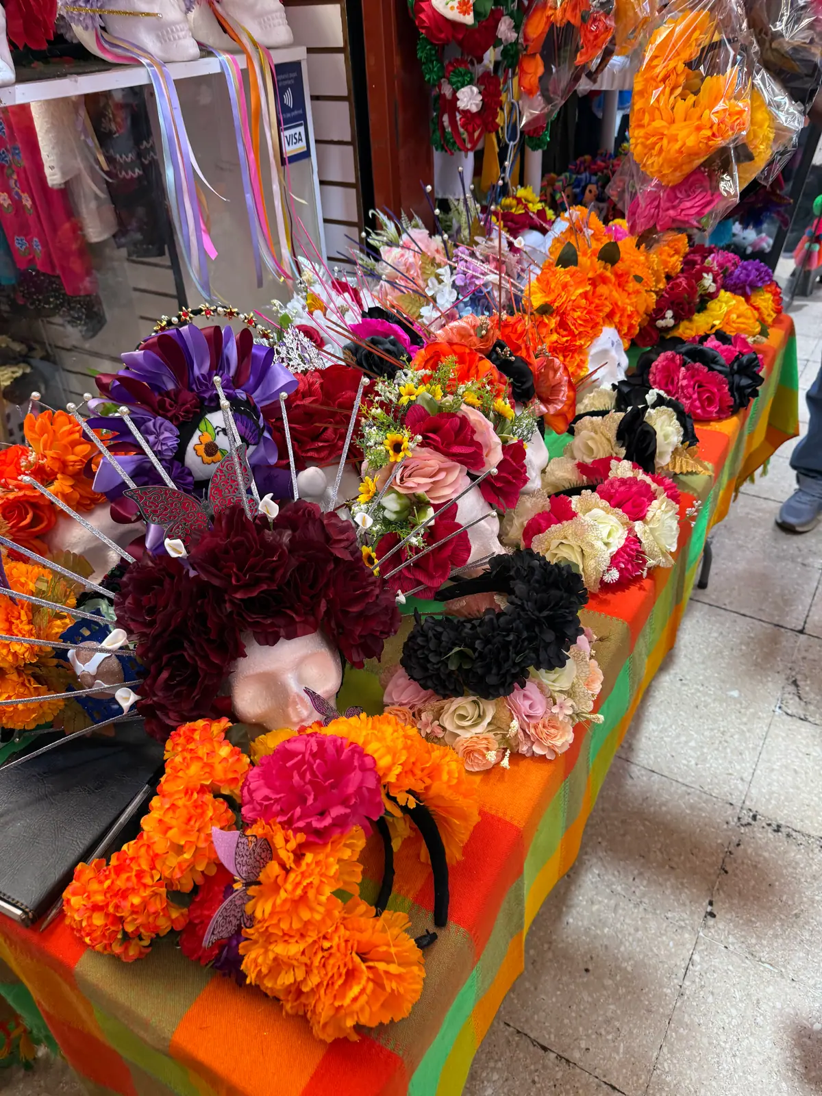 Coronas y diademas de flores para catrina, naranjas, rojas, moradas y negras, sobre una mesa con mantel de colores en el Mercado de Coyoacán