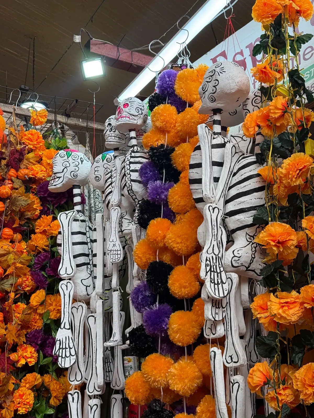 Esqueletos de cartón blancos colgados entre guirnaldas de flores naranjas, moradas y negras en un pasillo del Mercado de Coyoacán