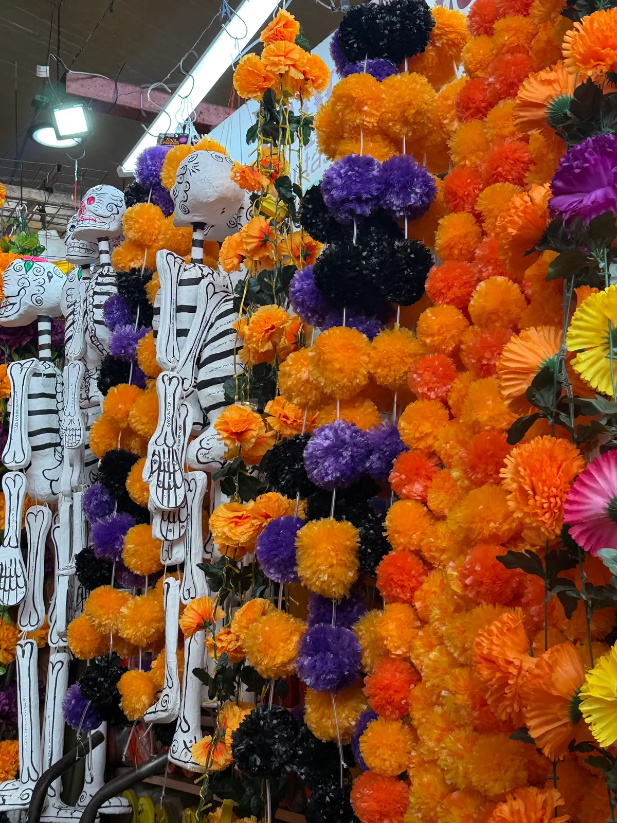 Guirnaldas de flores artificiales naranjas, moradas y negras colgadas junto a esqueletos de cartón blancos en un puesto del Mercado de Coyoacán