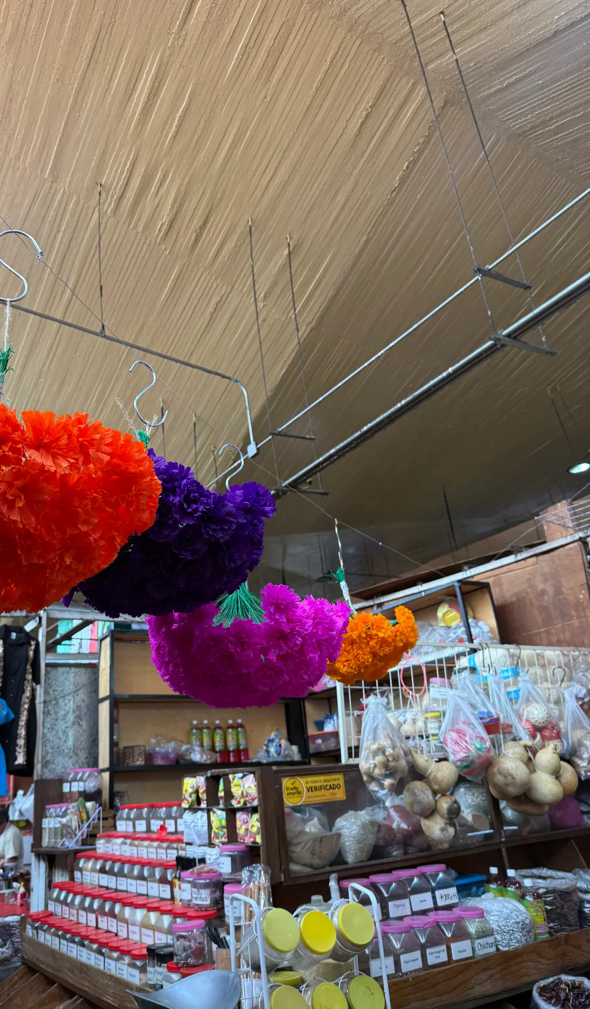 Flores grandes de papel naranja, morado y rosa colgadas sobre un puesto con frascos de semillas y frijoles, guajes y chiles secos