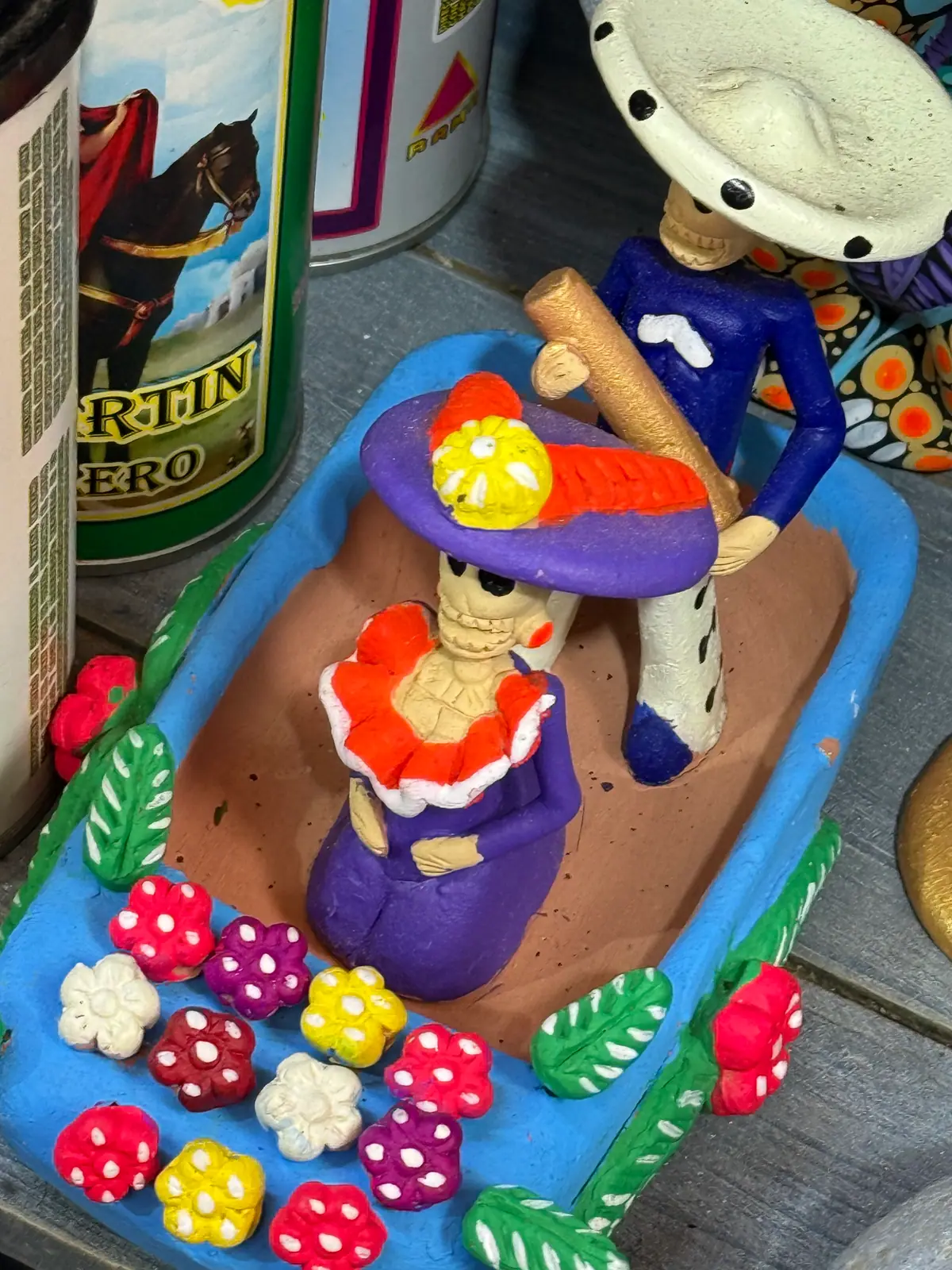Una trajinera de barro azul adornada con flores de colores, con una catrina de sombrero morado y un remero calavera