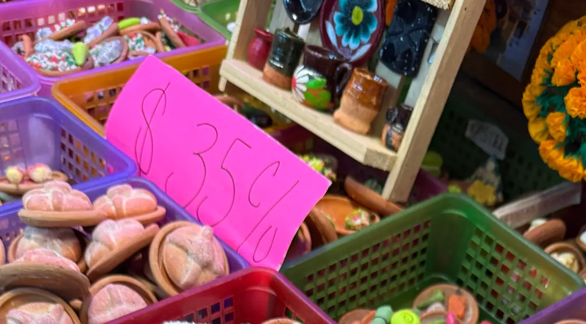 Panes de muerto en miniatura, de barro, sobre platitos, junto a un letrero rosa que dice 35 pesos cada uno y una repisa con jarritos de barro