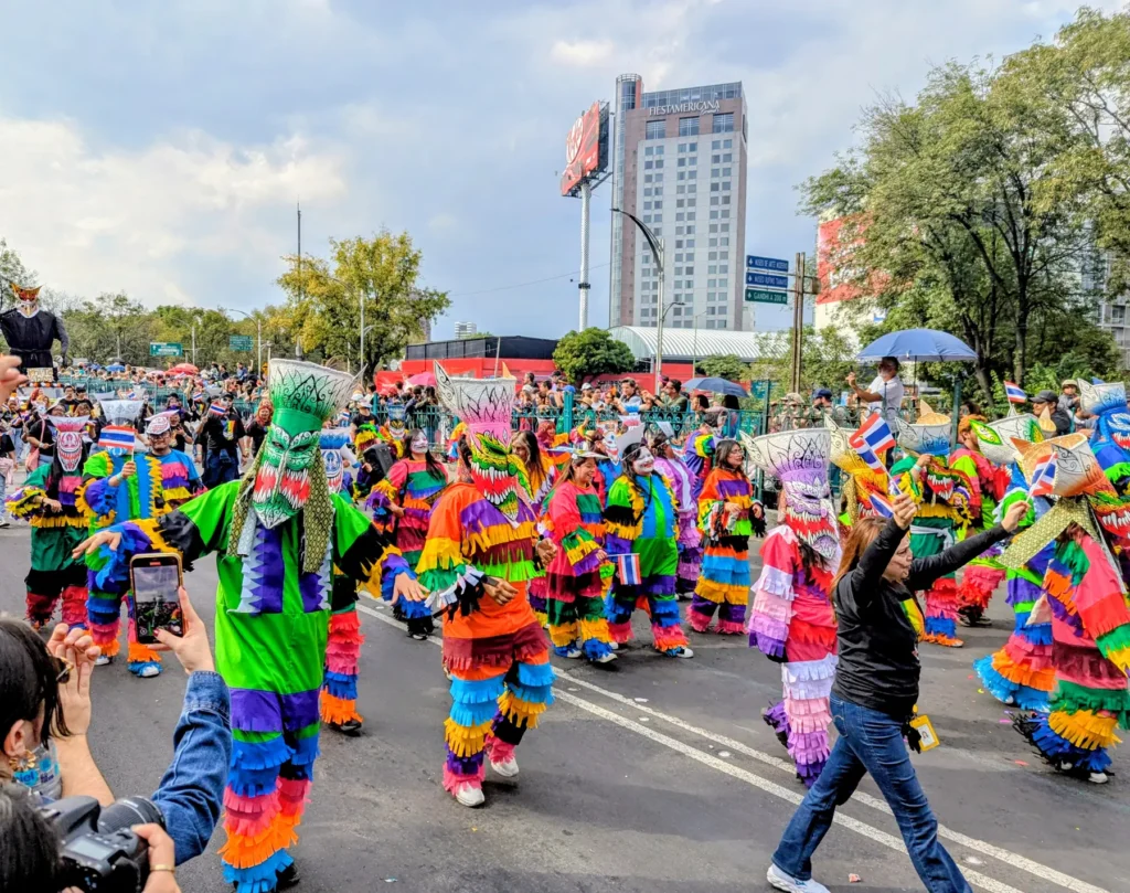 Una comparsa con trajes de flecos de colores baila sobre la avenida ante el público, en el Gran Desfile de Día de Muertos de 2025