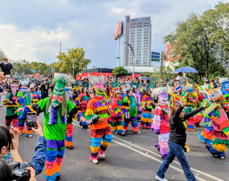 Una comparsa con trajes de flecos de colores baila sobre la avenida ante el público, en el Gran Desfile de Día de Muertos de 2025