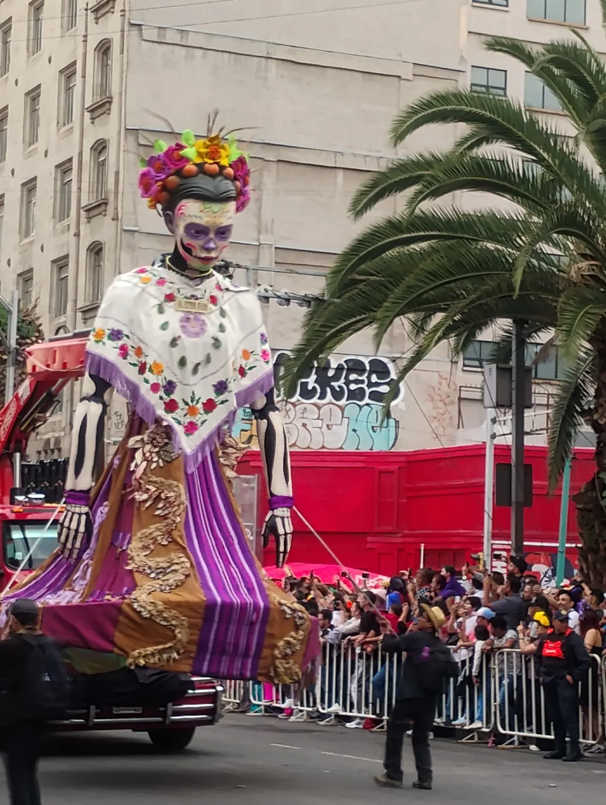 Una catrina monumental con mantilla blanca y corona de flores sobre un carro alegórico de fondo rojo, con el público detrás de la valla, en el Gran Desfile de 2025