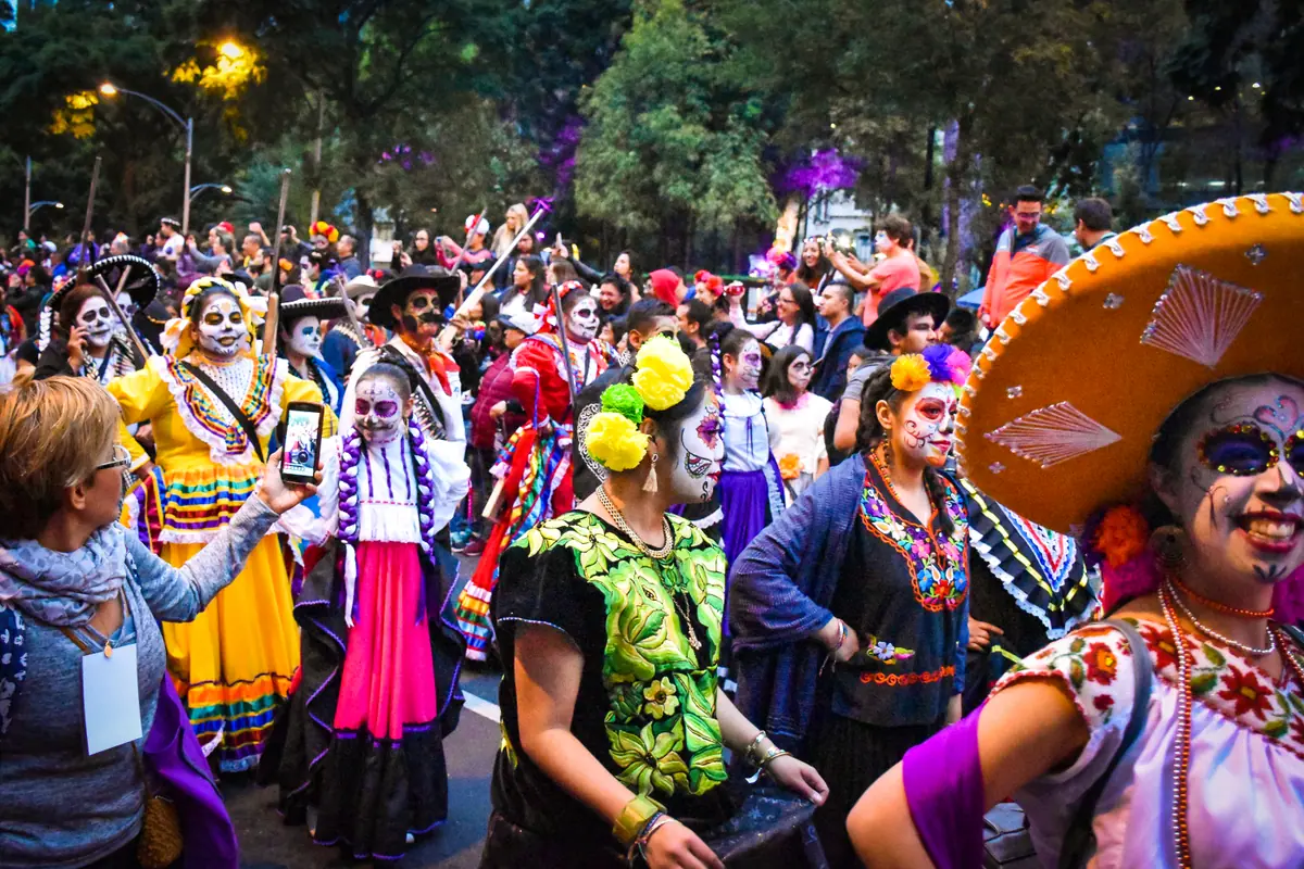 Contingentes de catrinas con sombreros de charro y trajes regionales avanzan al anochecer en la Mega Procesión de Catrinas
