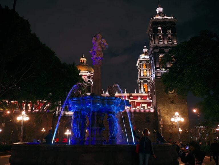 El Zócalo de Puebla de noche, con la fuente del Arcángel iluminada de azul y las torres de la Catedral encendidas
